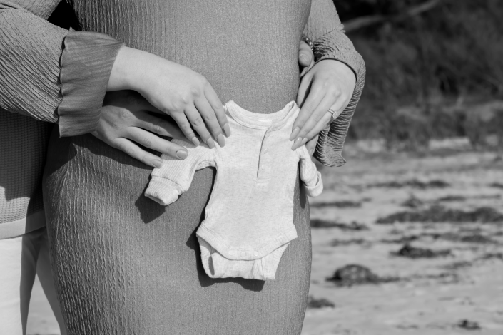 Playful moment of couple hugging and showing baby's bodysuit standing in wet sand, mother's dress flowing in wind, natural black and white joy captured