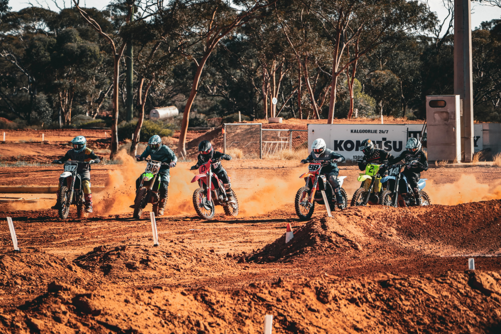 A group of motocross riders racing side by side on a dirt track, with spectators cheering in the background."