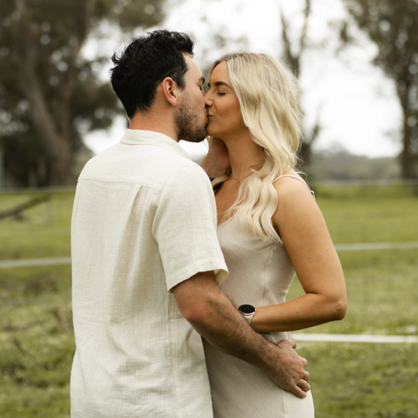 Husband kissing mother's while father embraces mother, white clothing creating ethereal effect against natural backdrop