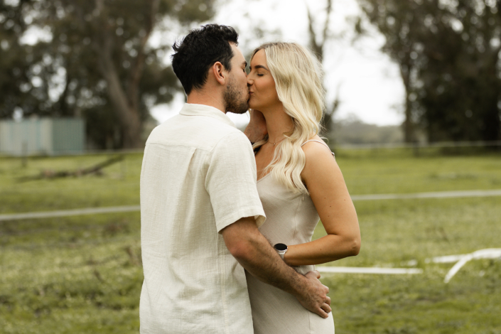 Husband kissing mother's while father embraces mother, white clothing creating ethereal effect against natural backdrop