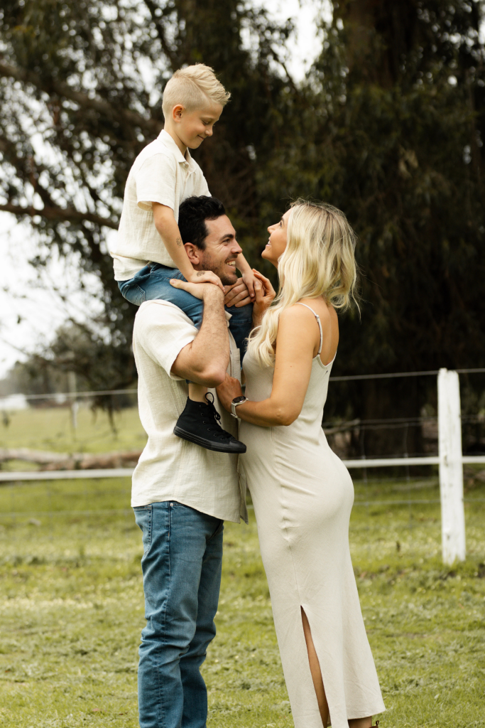Intimate profile of pregnant mother in white dress, father holding the young boy on his shoulders looking and smiling at her, backlit by soft sunset