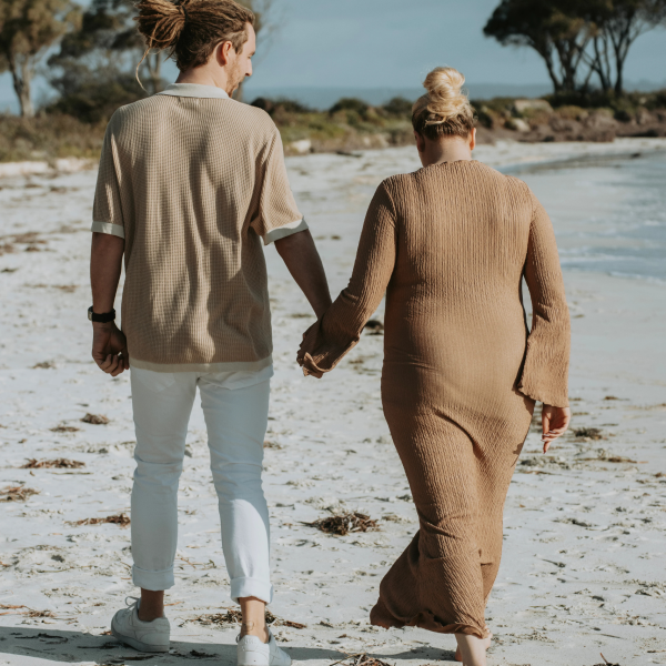 Expecting couple walking hand in hand along shoreline, soft pastel sky creating romantic atmosphere