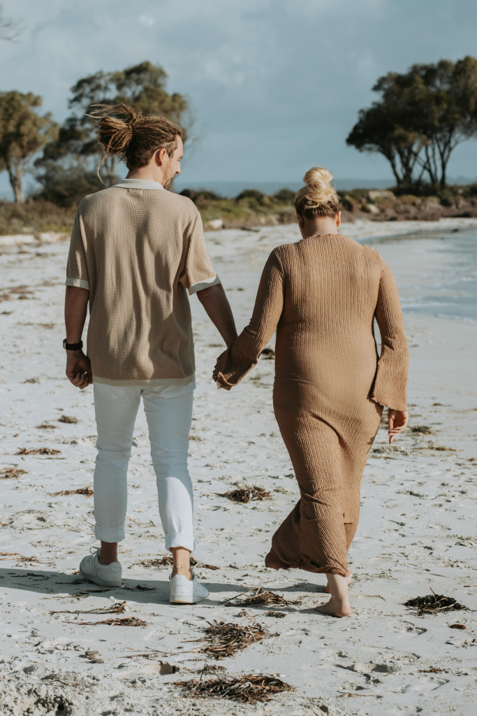 Expecting couple walking hand in hand along shoreline, soft pastel sky creating romantic atmosphere