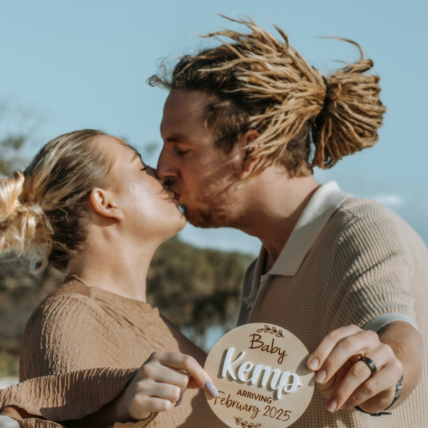 Couple sharing childs name, husband kissing wife while catching ocean breeze, golden light highlighting pregnancy glow