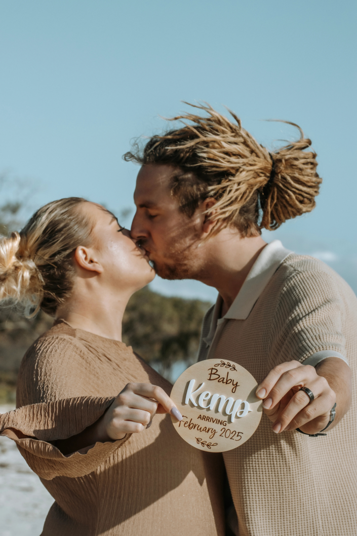 Couple sharing childs name, husband kissing wife while catching ocean breeze, golden light highlighting pregnancy glow