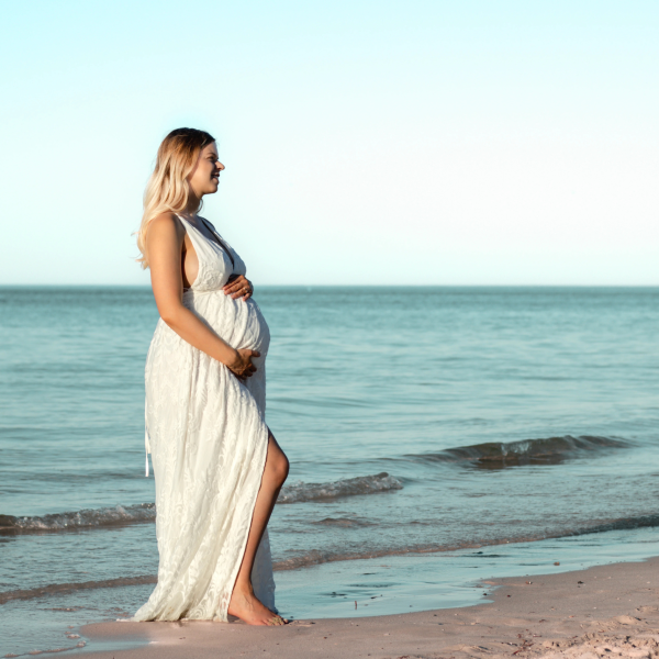Pregnant woman in a flowing white dress, enjoying a serene sunset at the beach, capturing the beauty of motherhood.