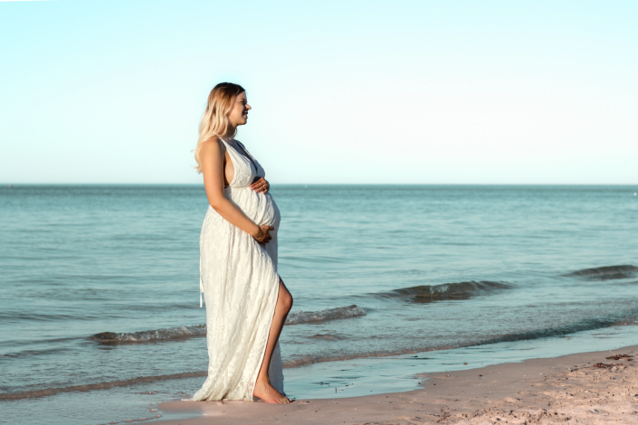 Pregnant woman in a flowing white dress, enjoying a serene sunset at the beach, capturing the beauty of motherhood.