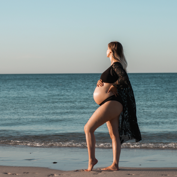 Expecting mother walking along the sandy shore, showcasing her baby bump against the backdrop of gentle ocean waves.