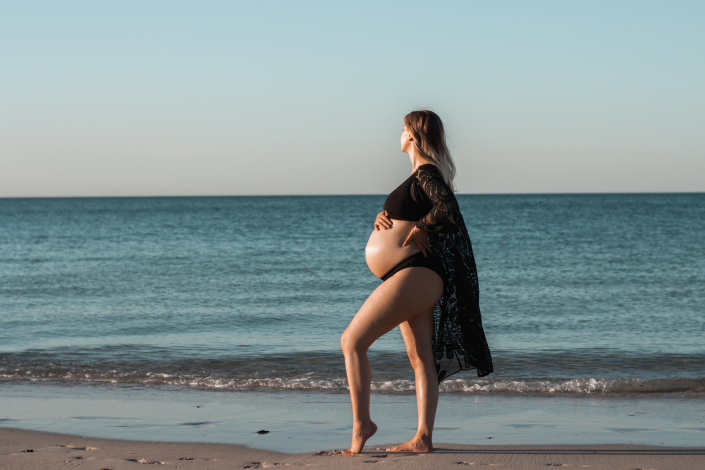 Expecting mother walking along the sandy shore, showcasing her baby bump against the backdrop of gentle ocean waves.