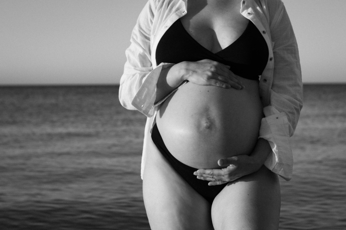 Beautiful pregnant woman relaxing on the beach, highlighting her baby bump with the sun setting over the horizon.