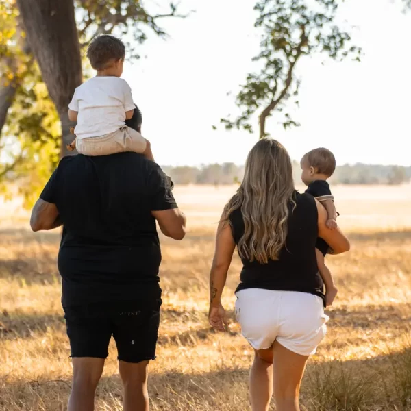 Candid family moment captured from behind, father carrying toddler on shoulders backlit by warm sunset, mother holding other child's hand as they walk through pastoral landscape