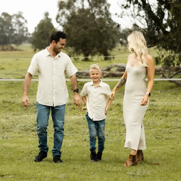 Ethereal maternity family portrait, parents and young child in flowing white outfits walking hand-in-hand through sun-drenched farm field, mother's pregnancy beautifully silhouetted against soft natural light