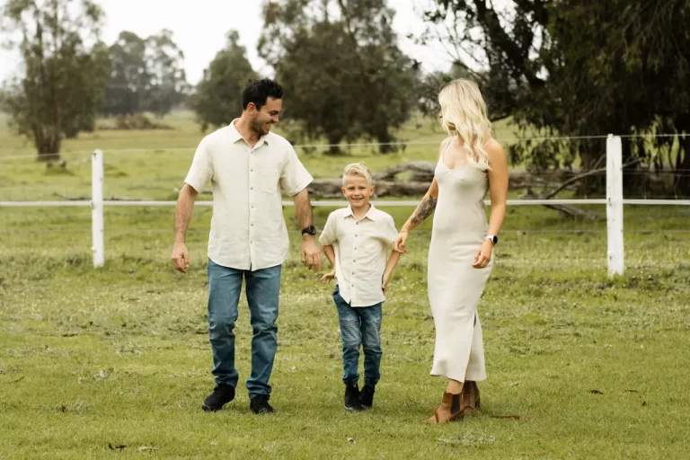 Ethereal maternity family portrait, parents and young child in flowing white outfits walking hand-in-hand through sun-drenched farm field, mother's pregnancy beautifully silhouetted against soft natural light
