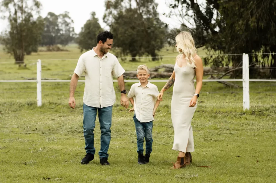 Ethereal maternity family portrait, parents and young child in flowing white outfits walking hand-in-hand through sun-drenched farm field, mother's pregnancy beautifully silhouetted against soft natural light
